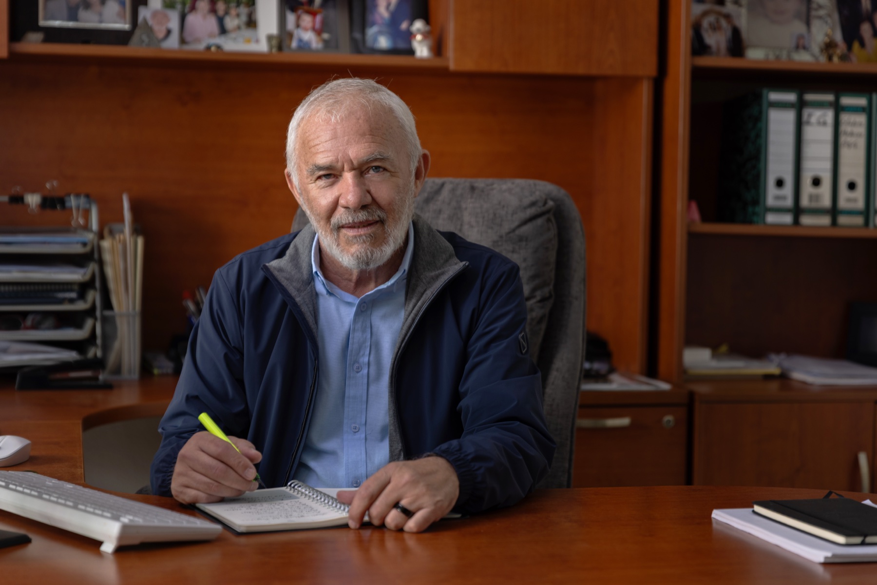 Directivo de cabello y barba blancos escribiendo en su libreta, en su oficina de madera con portarretratos familiares al fondo.