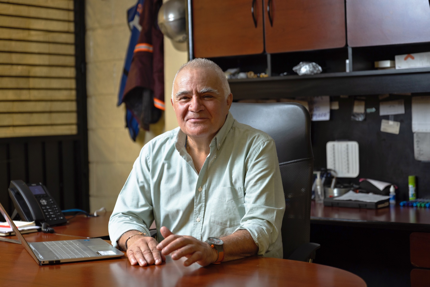 Mismo directivo en su oficina de planta, sentado al escritorio con laptop y teléfono.