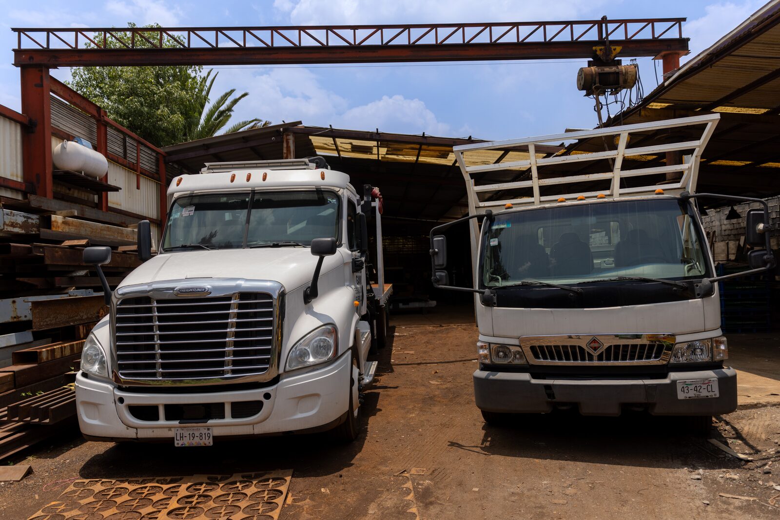 El tractocamión y un camión de redilas International estacionados bajo el puente grúa rojo del patio.