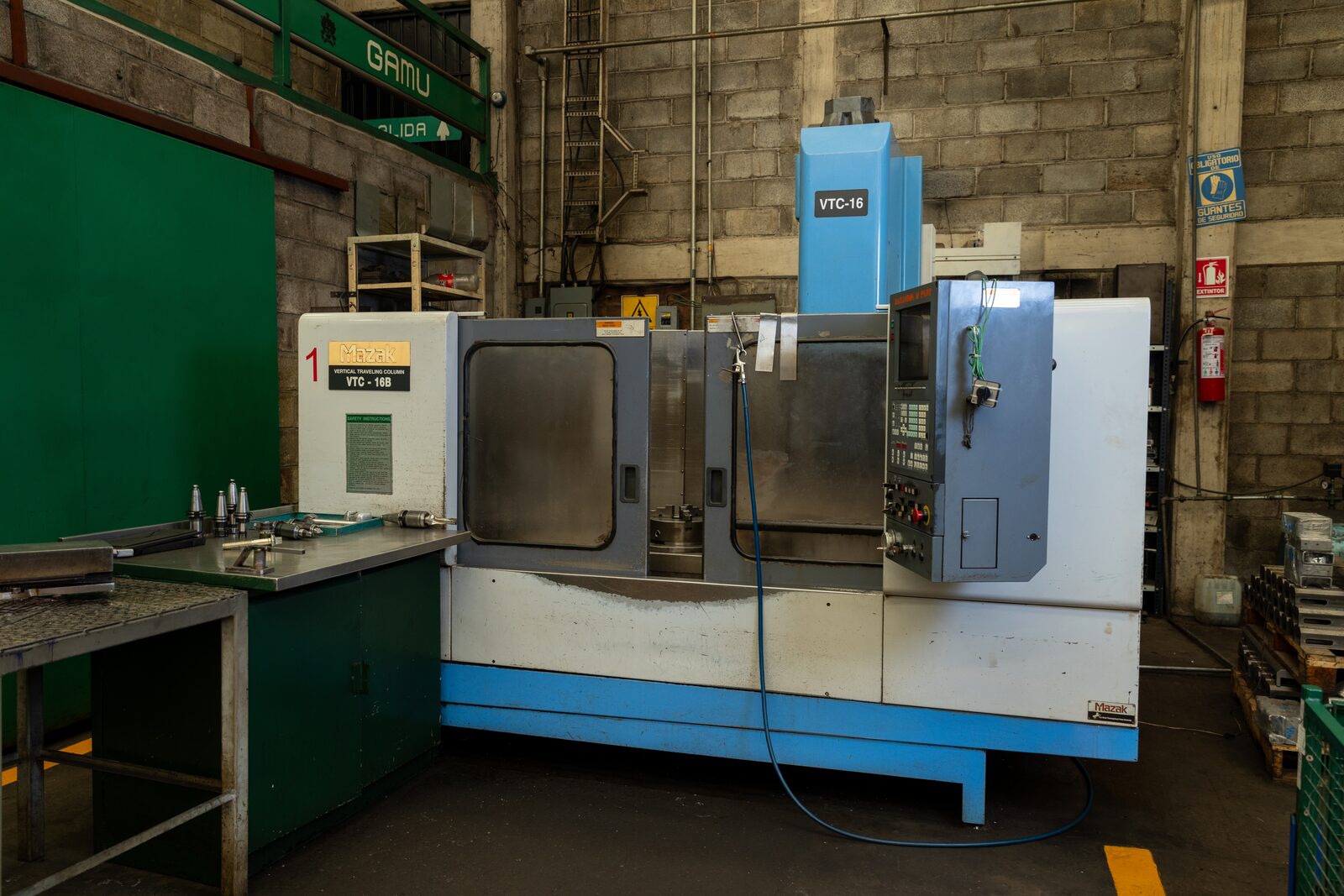 Mazak VTC-16B (nº 1) con portaherramientas alineados en el banco; letrero verde GAMU en el muro.